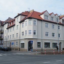 Mat-Bud Sp. z o.o. - Odnowiona kamienica narożna z czerwonym dachem, jasną elewacją i białymi oknami, widoczna z perspektywy ulicy w słoneczny dzień.