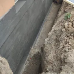 Świeżo zaizolowany fundament budynku z widoczną warstwą izolacyjną i wykopem ochronnym, zabezpieczonym przezroczystym zadaszeniem.