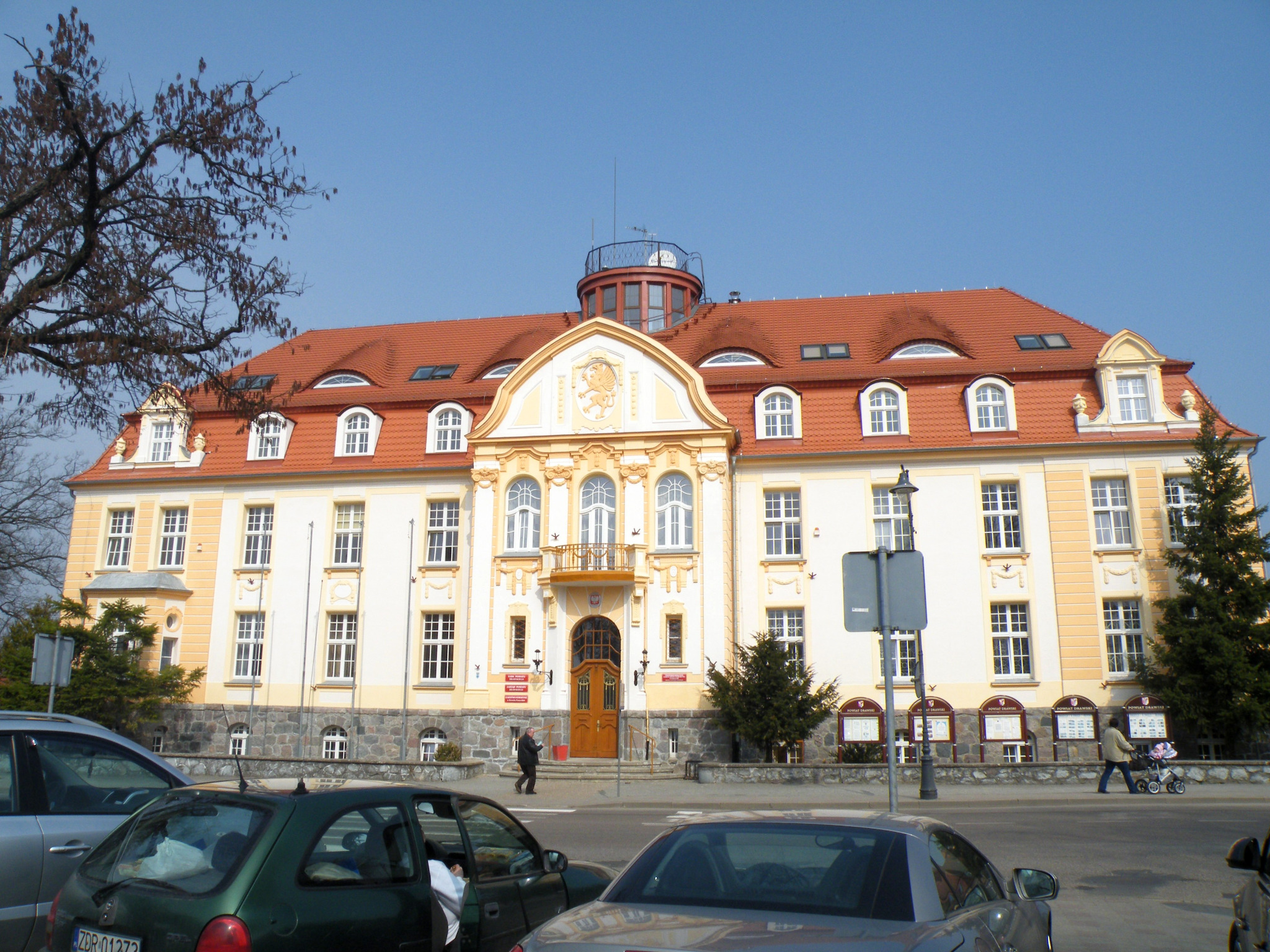 Elewacja odnowionego, trzypiętrowego budynku publicznego z pomarańczowym dachem, widoczna zza zaparkowanych samochodów.