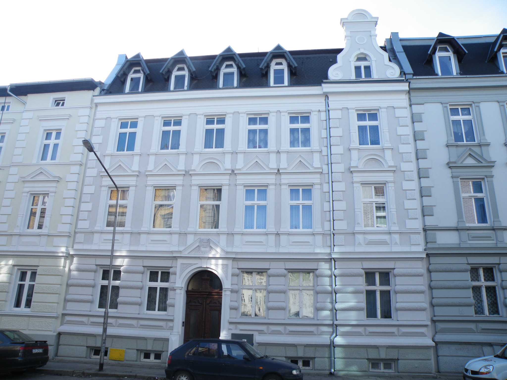 Odnowiona fasada kamienicy z ozdobnymi elementami architektonicznymi, widoczne okna z firankami, ciemny dach z lukarnami, ulica z zaparkowanymi samochodami.