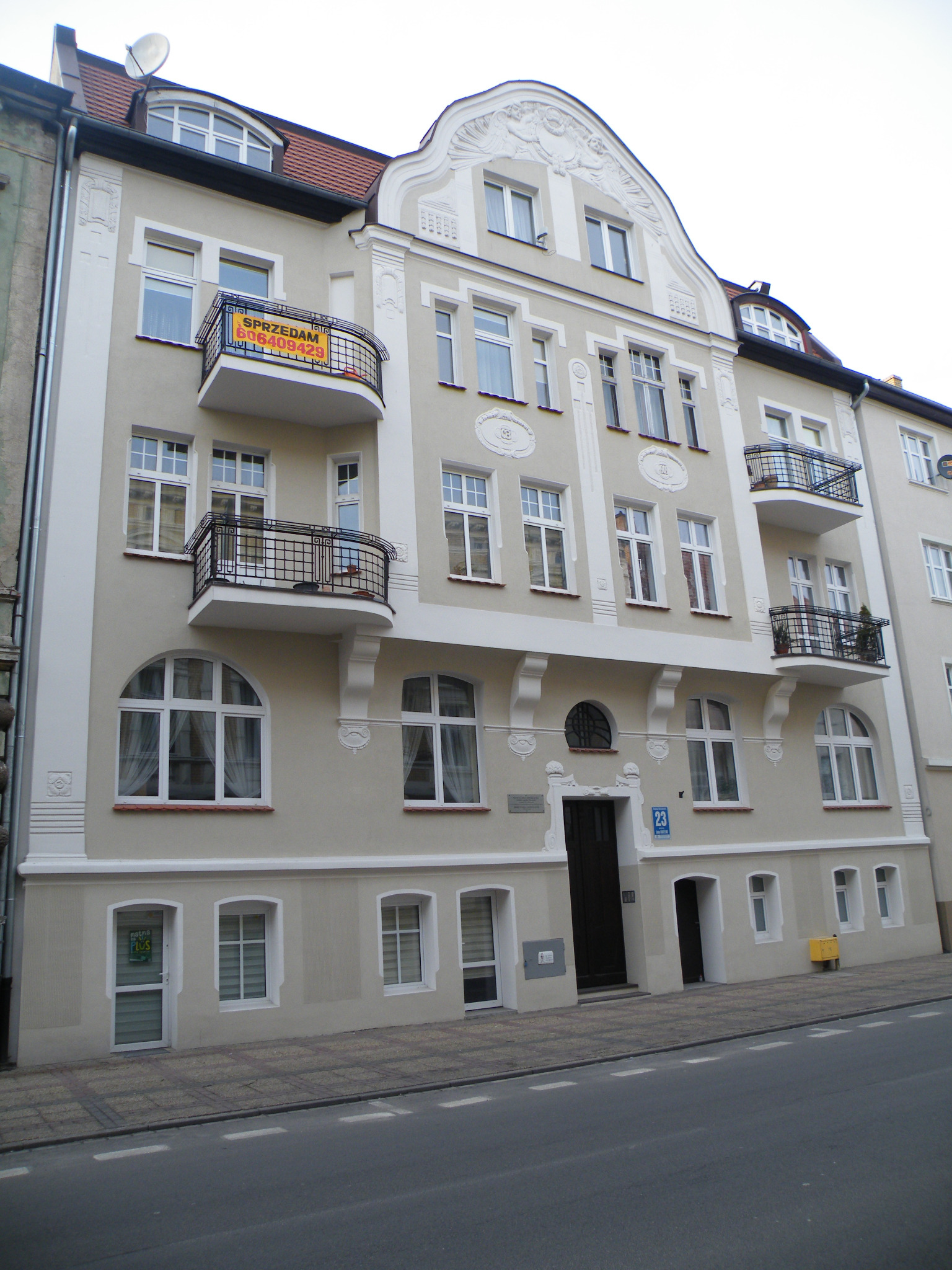 Odnowiona kamienica z beżową elewacją, białymi zdobieniami i balkonami z czarną balustradą. Na jednym z balkonów widoczna tabliczka 'Sprzedam'.