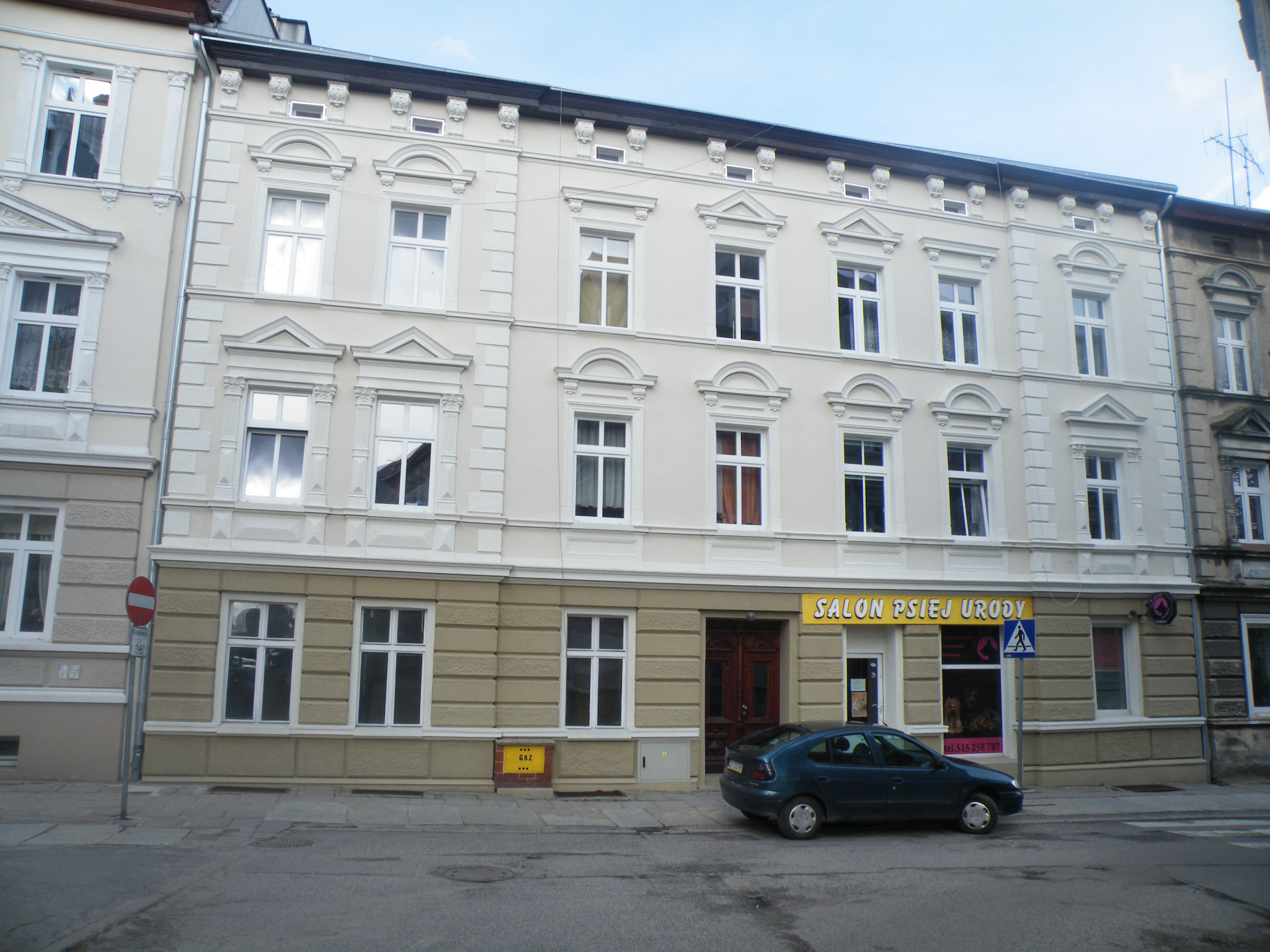 Odnowiona fasada trzypiętrowej kamienicy z ozdobnymi elementami architektonicznymi, widok z poziomu ulicy, zaparkowany samochód i szyld salonu usługowego.