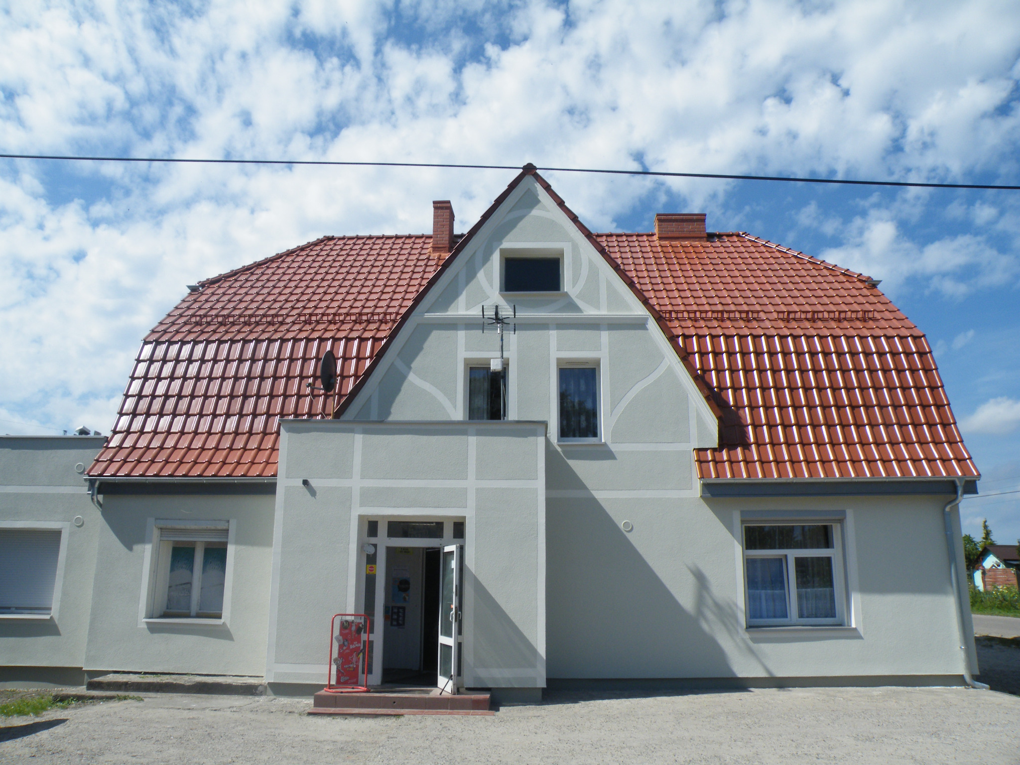 Odnowiony budynek z czerwoną dachówką karpiówką i jasnoszarą elewacją w słoneczny dzień z błękitnym niebem i chmurami.