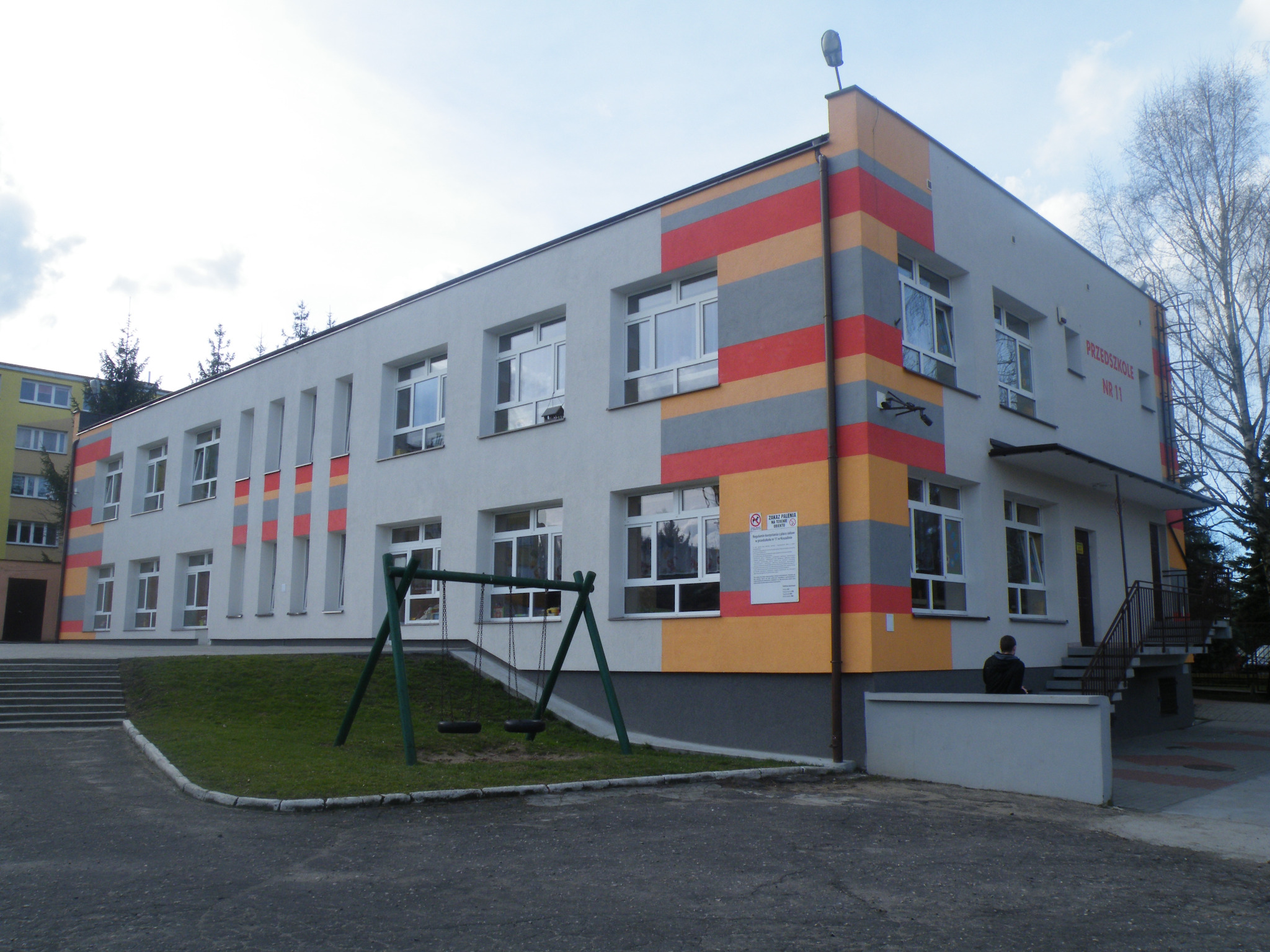 Budynek przedszkola z pomalowaną elewacją w pasy szaro-pomarańczowo-czerwone, widoczne okna, huśtawka i schody wejściowe.