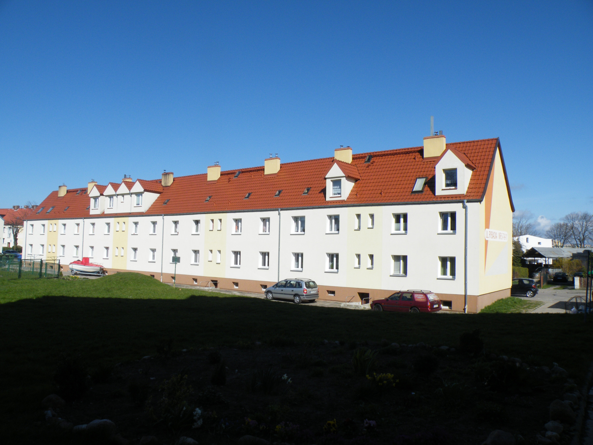 Długi, trzypiętrowy budynek mieszkalny z czerwonym dachem, widoczny z perspektywy trawnika, z zaparkowanymi samochodami i łodzią na przyczepie w tle.