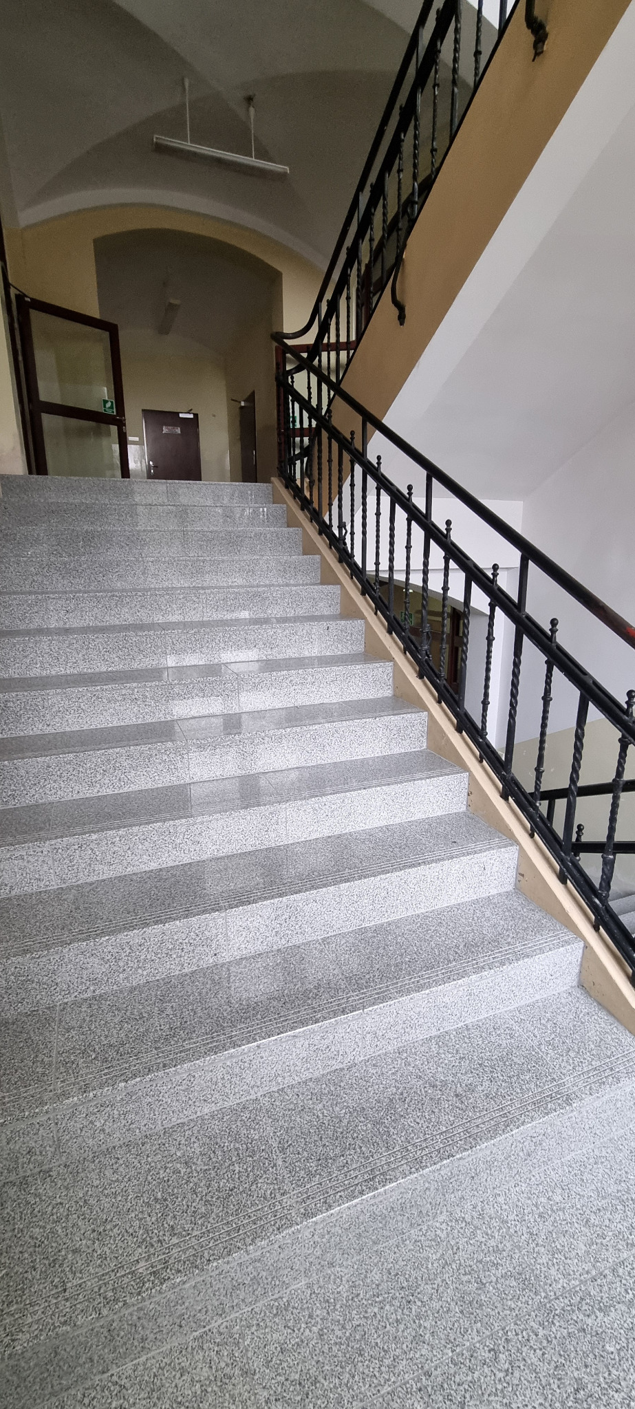 Granitowe schody z czarną, kutą balustradą w budynku z beżowymi ścianami i łukowym przejściem w tle.