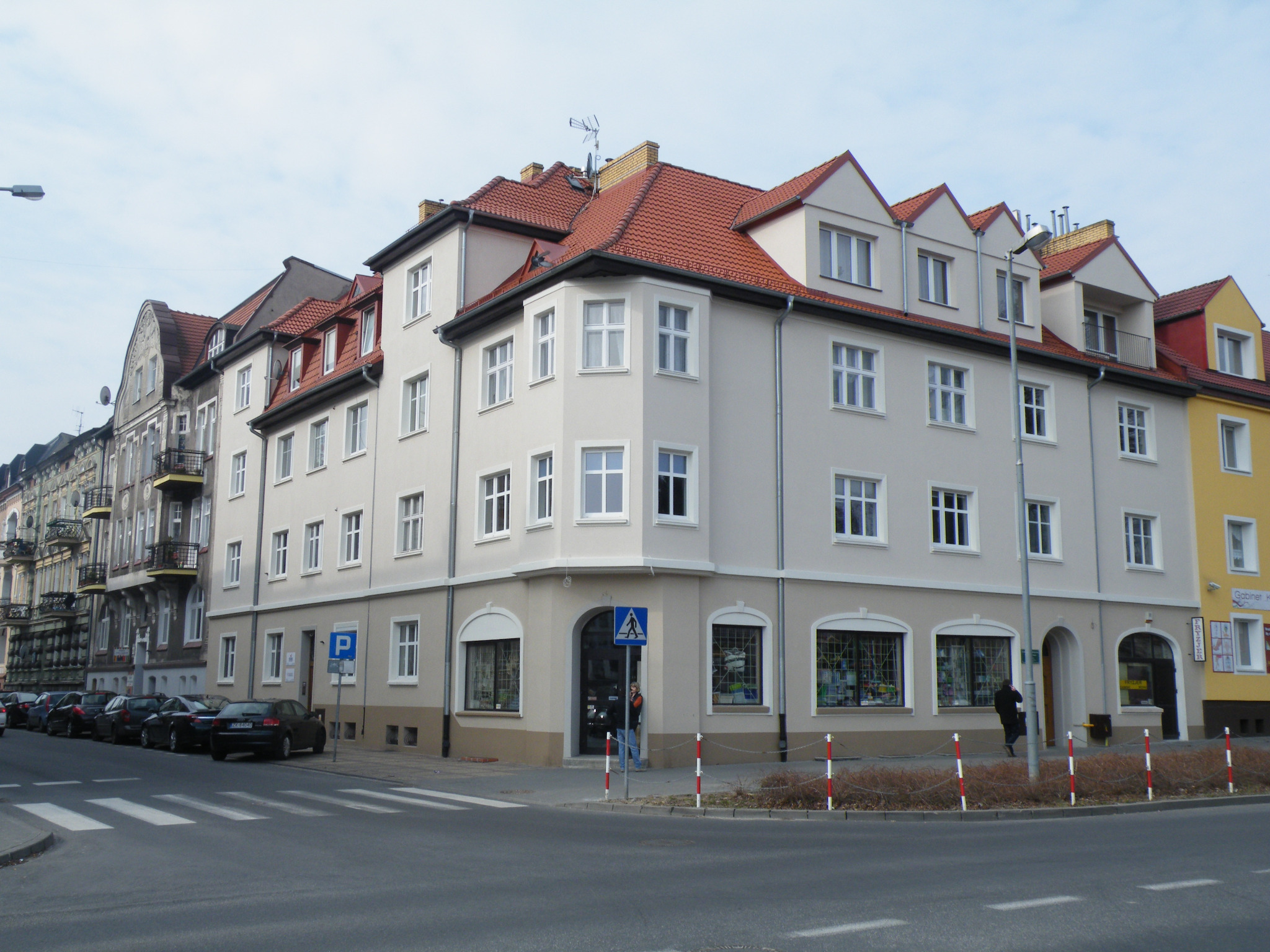 Odnowiona kamienica narożna z czerwonym dachem, jasną elewacją i białymi oknami, widoczna z perspektywy ulicy w słoneczny dzień.