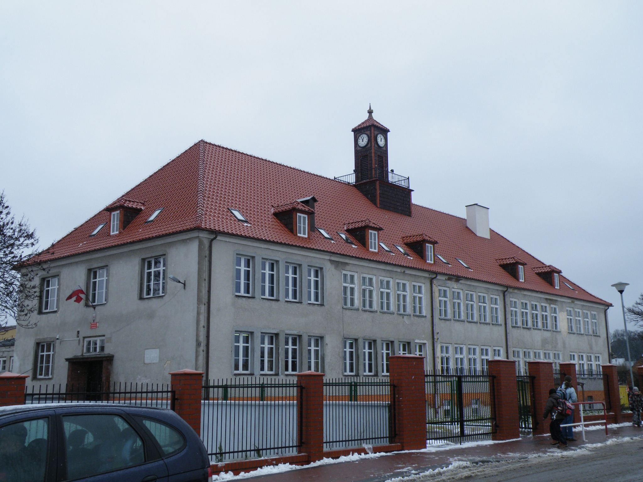 Duży, dwupiętrowy budynek z czerwonym dachem i wieżą zegarową, widoczny zza metalowego ogrodzenia z ceglanymi słupkami, częściowo zasłonięty przez samochód na pierwszym planie.