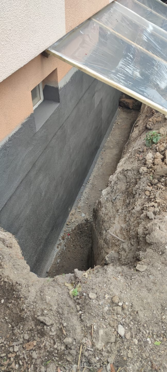 Świeżo zaizolowany fundament budynku z widoczną warstwą izolacyjną i wykopem ochronnym, zabezpieczonym przezroczystym zadaszeniem.