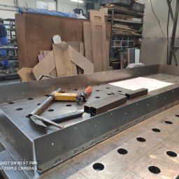 Roman Długosz OZE - Metalowa konstrukcja na perforowanym stole spawalniczym z narzędziami i rysunkiem technicznym, spawacz w tle. Detal z procesu obróbki metalu.