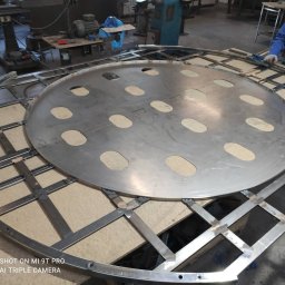 Roman Długosz OZE - Konstrukcja metalowa z okrągłą płytą z otworami, spawana przez spawacza. Widoczne spawy i elementy konstrukcyjne.