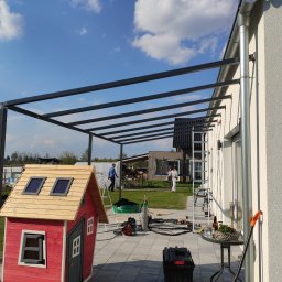 Roman Długosz OZE - Montaż metalowej pergoli przy domu jednorodzinnym. Widoczne narzędzia, butla z gazem i drabina. W tle domek dla dzieci i osoby na posesji.