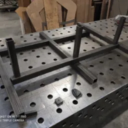 Stalowa konstrukcja z profili kwadratowych na perforowanym stole spawalniczym, widoczne narzędzia i elementy, przygotowana do spawania.