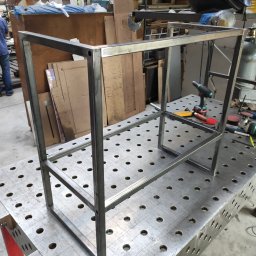 Roman Długosz OZE - Metalowa konstrukcja spawana na stole spawalniczym z narzędziami, widoczny fragment warsztatu i sylwetka osoby w tle. Konstrukcja w formie prostopadłościanu.