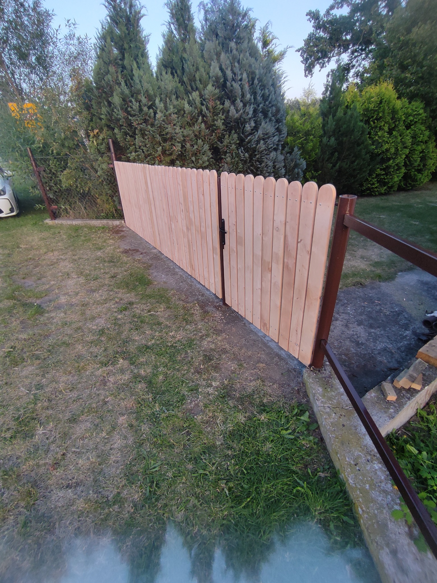 Nowy, jasny, drewniany płot z furtką, z zaokrąglonymi deskami, zamontowany na metalowych słupkach, na tle zielonego ogrodu z drzewami.