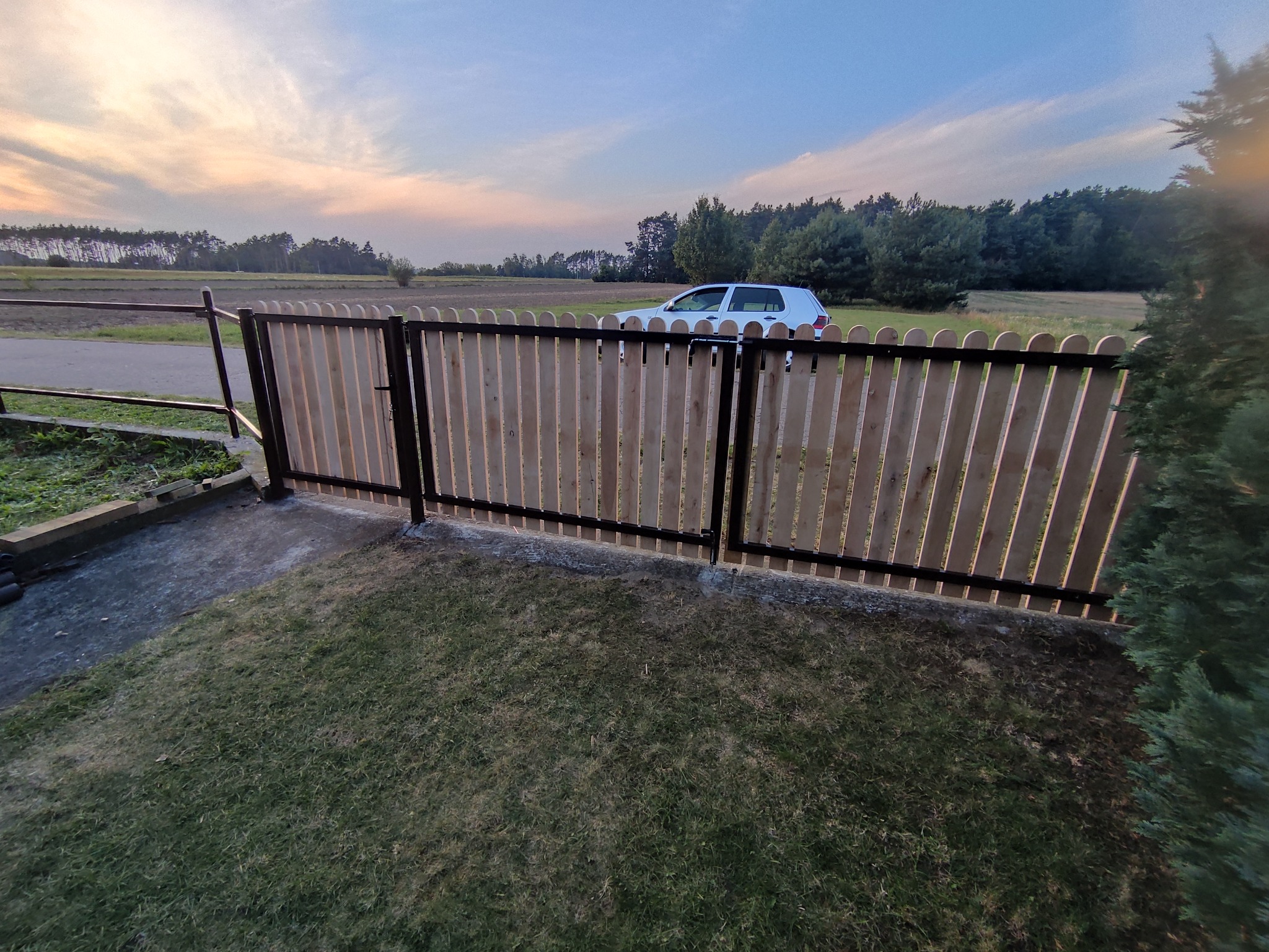 Nowoczesne ogrodzenie drewniane z czarnymi elementami stalowymi, brama zamknięta, w tle samochód i zachodzące słońce. Estetyczne wykończenie posesji.