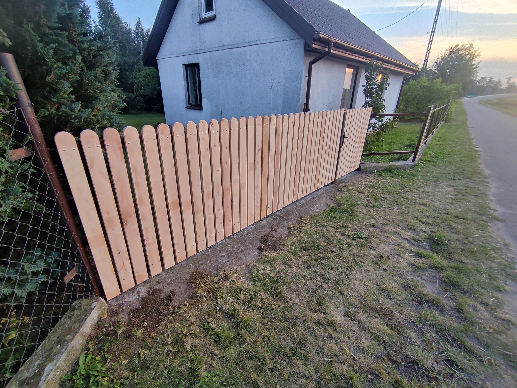 Nowy drewniany płot z furtką przed domem. Płot z pionowych desek z zaokrąglonymi górnymi krawędziami, przymocowanych na metalowych słupkach. Widok na dom i drogę.
