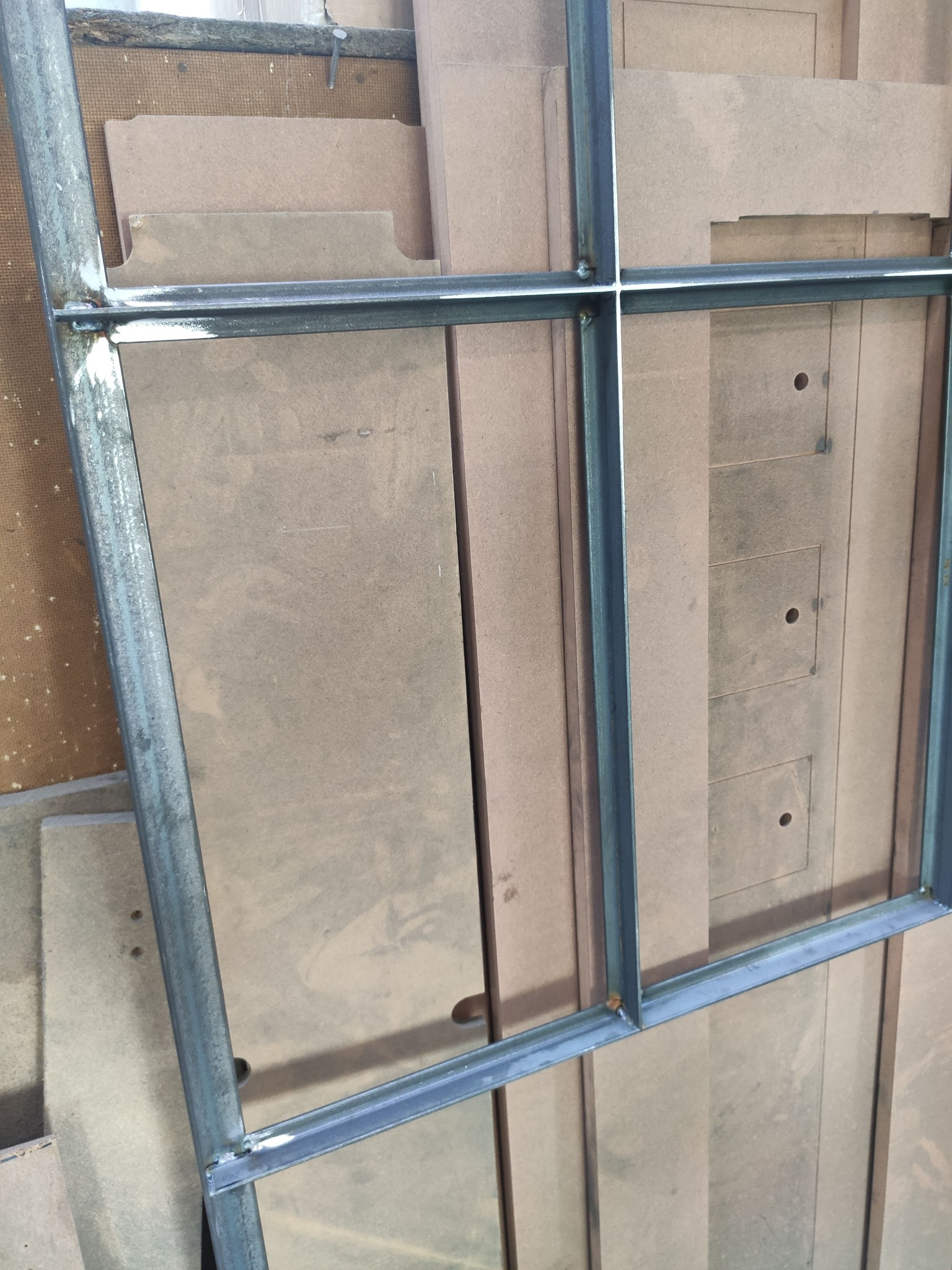 Spawana rama z metalowych profili na tle płyt MDF. Widoczne spawy i surowe wykończenie metalu. Konstrukcja podzielona na kwadratowe pola.