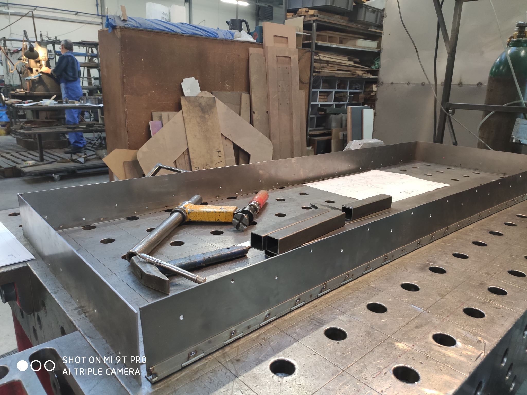 Metalowa konstrukcja na perforowanym stole spawalniczym z narzędziami i rysunkiem technicznym, spawacz w tle. Detal z procesu obróbki metalu.