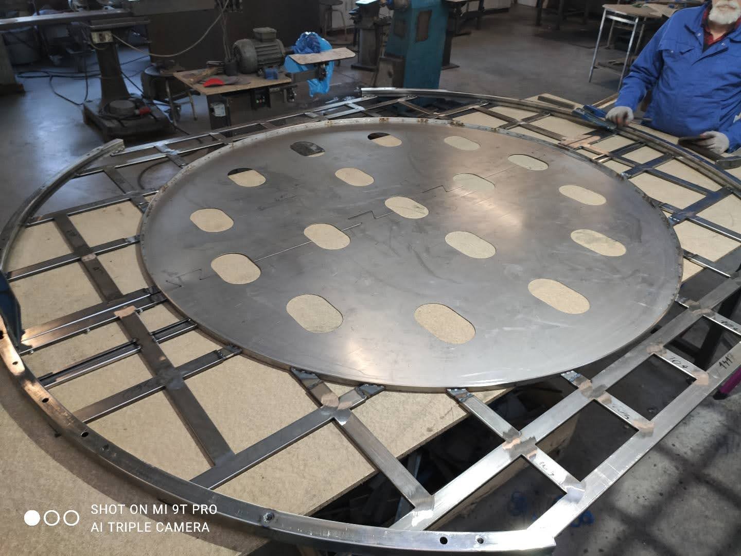 Konstrukcja metalowa z okrągłą płytą z otworami, spawana przez spawacza. Widoczne spawy i elementy konstrukcyjne.