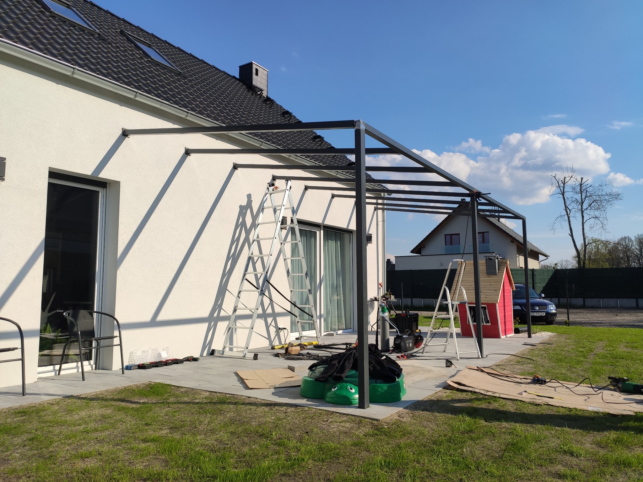 Montaż metalowej konstrukcji zadaszenia tarasu przy białym domu jednorodzinnym. Widoczne narzędzia i drabiny. Błękitne niebo w tle.