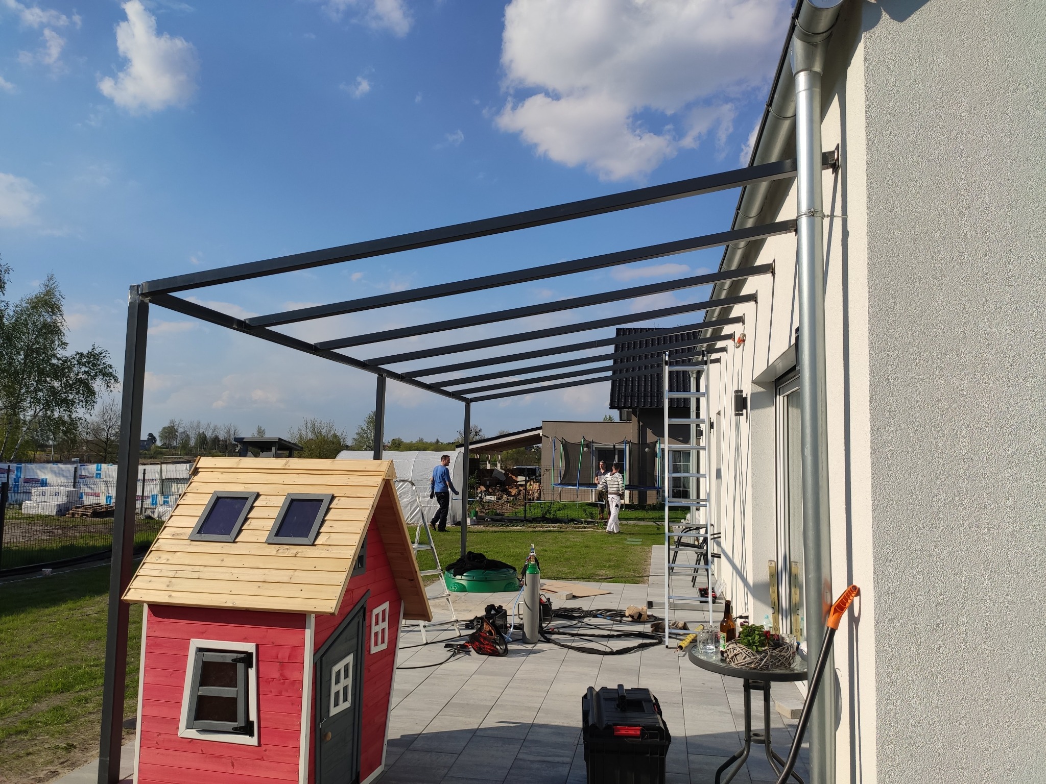 Montaż metalowej pergoli przy domu jednorodzinnym. Widoczne narzędzia, butla z gazem i drabina. W tle domek dla dzieci i osoby na posesji.