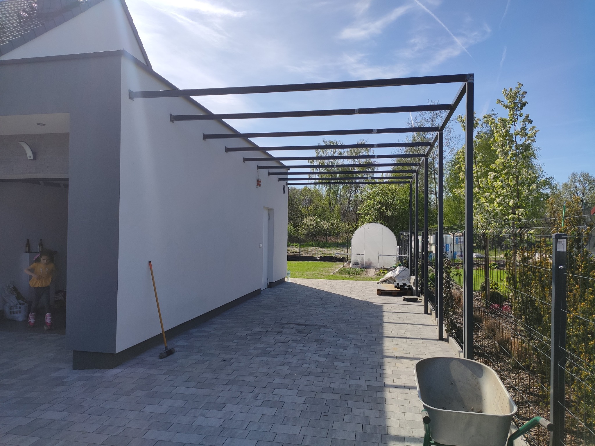 Metalowa pergola przy białym domu z widokiem na ogród i szklarnię, błękitne niebo w tle. Widoczny fragment kostki brukowej i taczka.