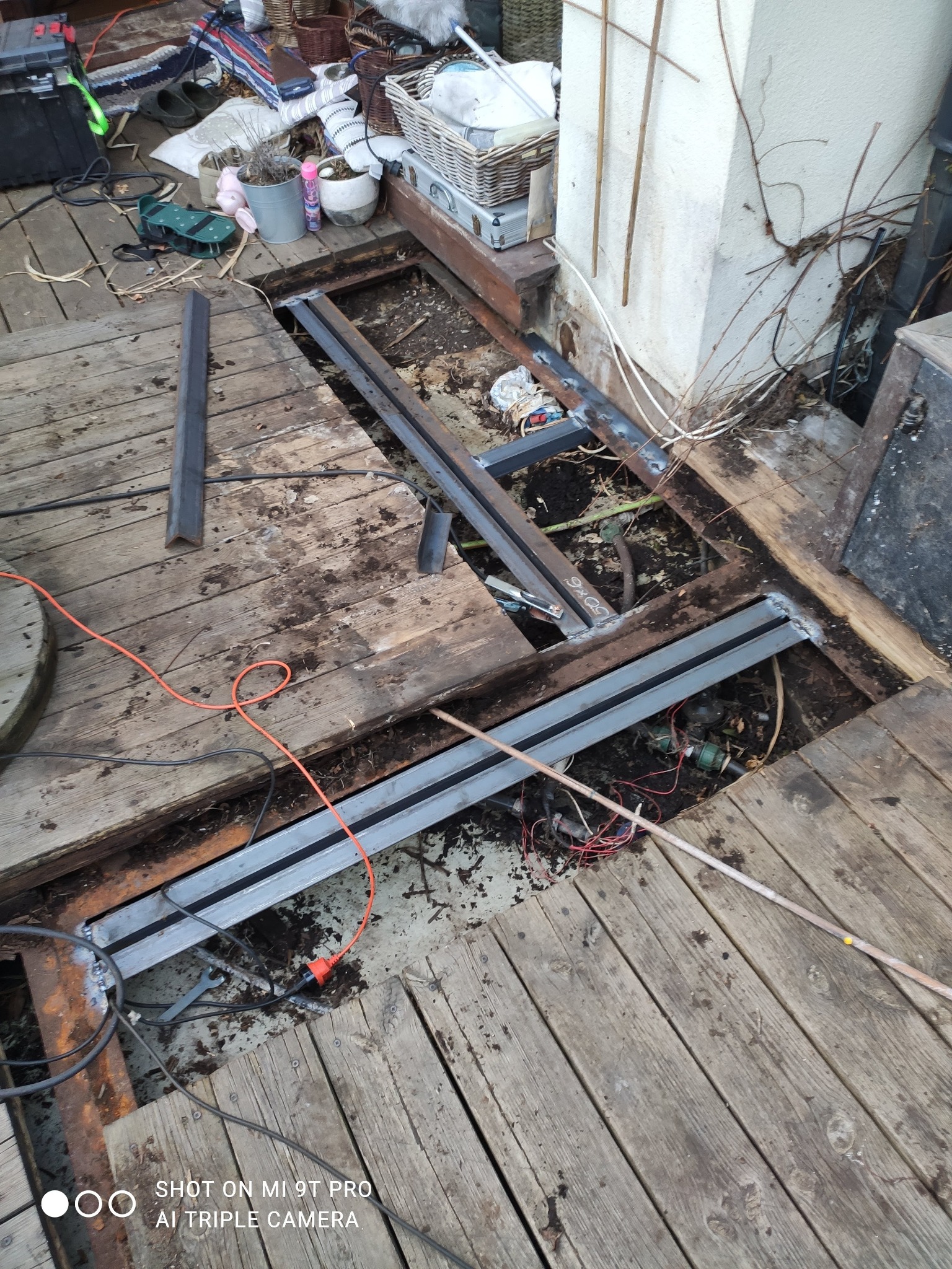 Metalowa konstrukcja spawana w drewnianej podłodze tarasu, widoczne narzędzia i przewody. Etap prac spawalniczych w przestrzeni zewnętrznej.