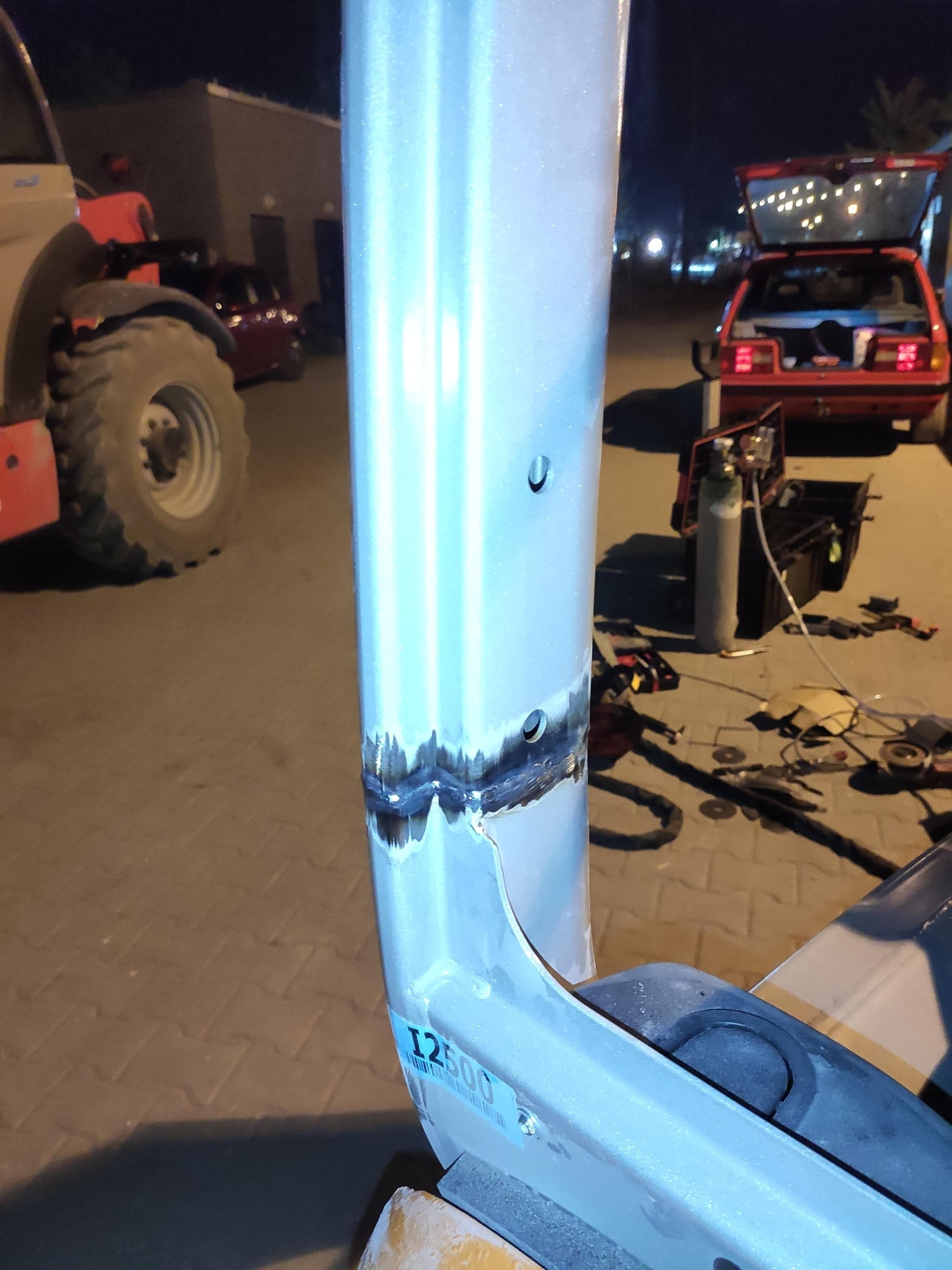 Spoina na metalowym elemencie maszyny budowlanej, widoczne ślady spawania i pobliskie narzędzia na zewnątrz, w tle czerwony samochód z otwartym bagażnikiem.