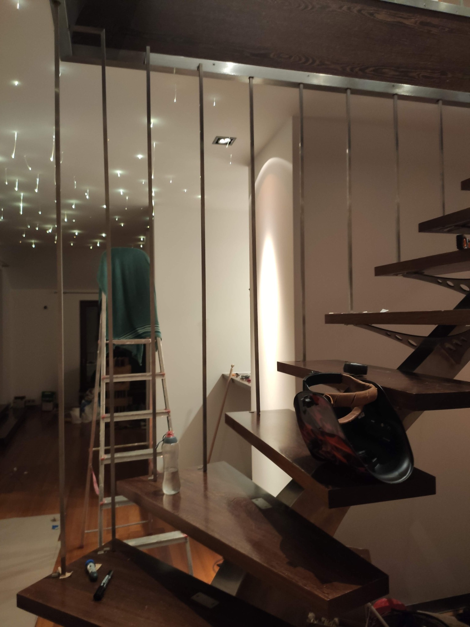 Metalowe barierki schodowe w trakcie montażu, widoczne narzędzia pracy i drabina. Oświetlenie sufitowe w tle, schody drewniane. Prace wykończeniowe.