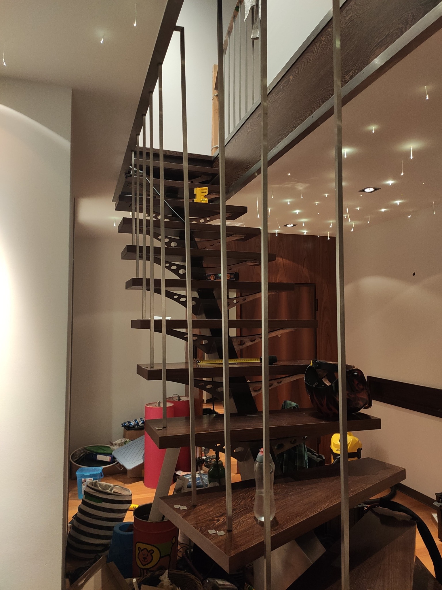 Nowoczesne, drewniane schody wiszące z metalowymi barierkami w jasnym wnętrzu. Widoczne oświetlenie sufitowe i elementy wykończenia.