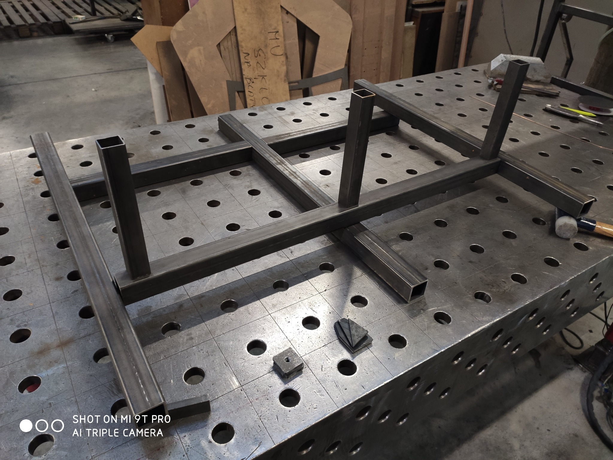 Stalowa konstrukcja z profili kwadratowych na perforowanym stole spawalniczym, widoczne narzędzia i elementy, przygotowana do spawania.