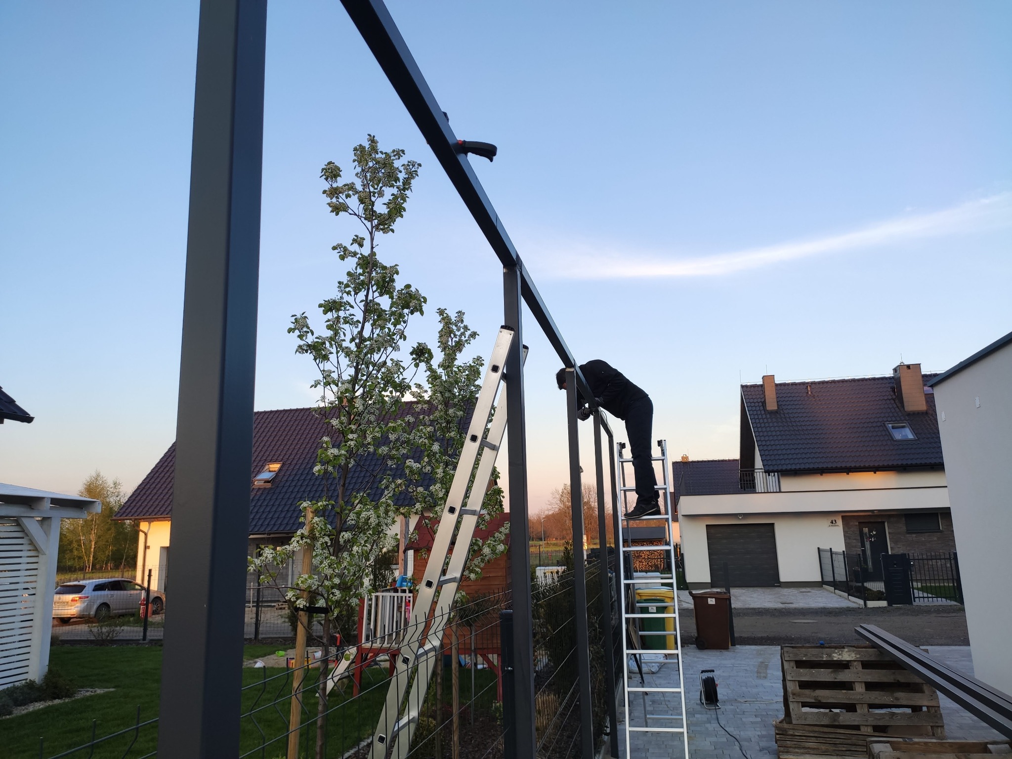 Montaż metalowej konstrukcji ogrodzenia przez pracownika na drabinie, z widocznymi elementami spawania i narzędziami, na tle domów i zieleni.