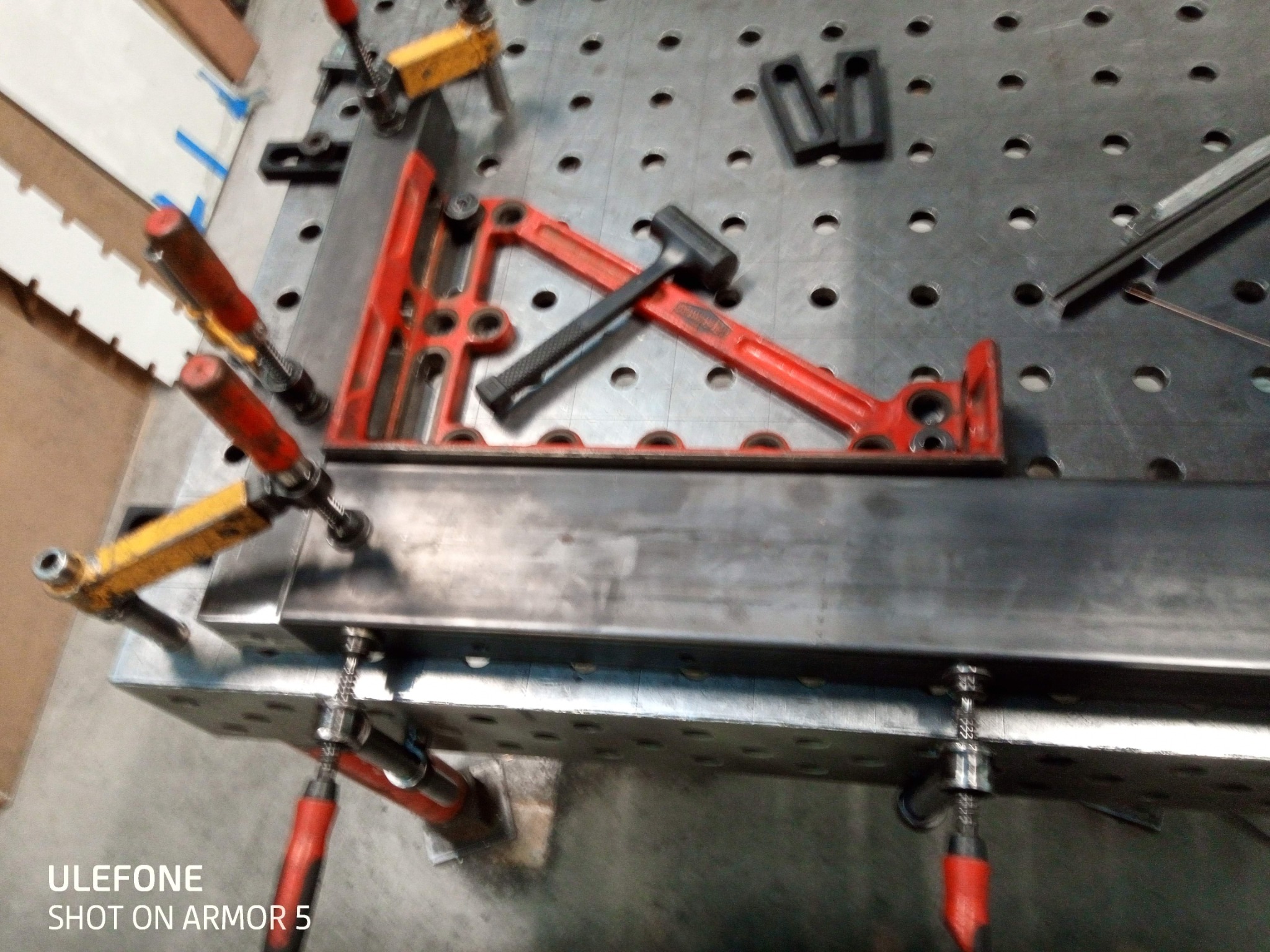 Metalowa konstrukcja spawana na perforowanym stole z użyciem kątownika, młotka i ścisków. Detal precyzyjnego łączenia elementów pod kątem prostym.