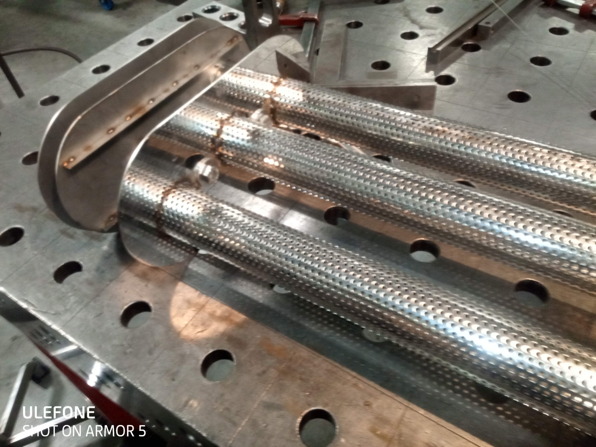 Metalowa konstrukcja z perforowanych rur na stole spawalniczym, widoczne ślady spawania i uchwyty. Obróbka metalu, precyzyjne łączenie elementów.