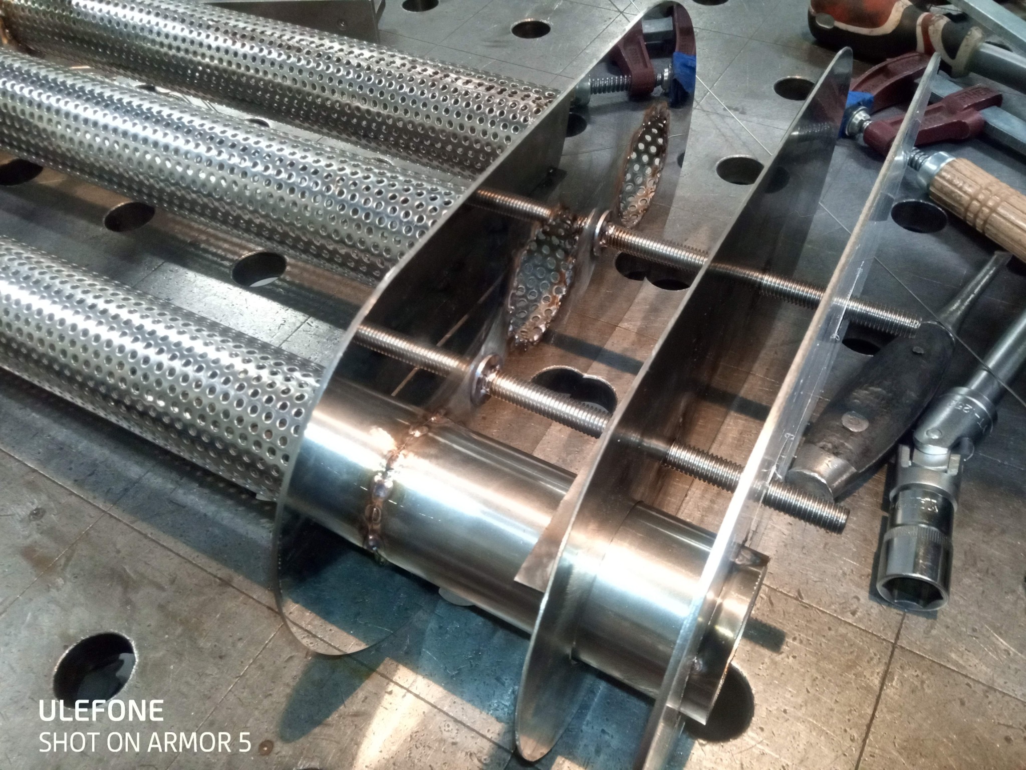 Metalowa konstrukcja z perforowanych rur i płaskich elementów, połączona śrubami, widoczna na stole warsztatowym z narzędziami. Detal spawalniczy na rurze.