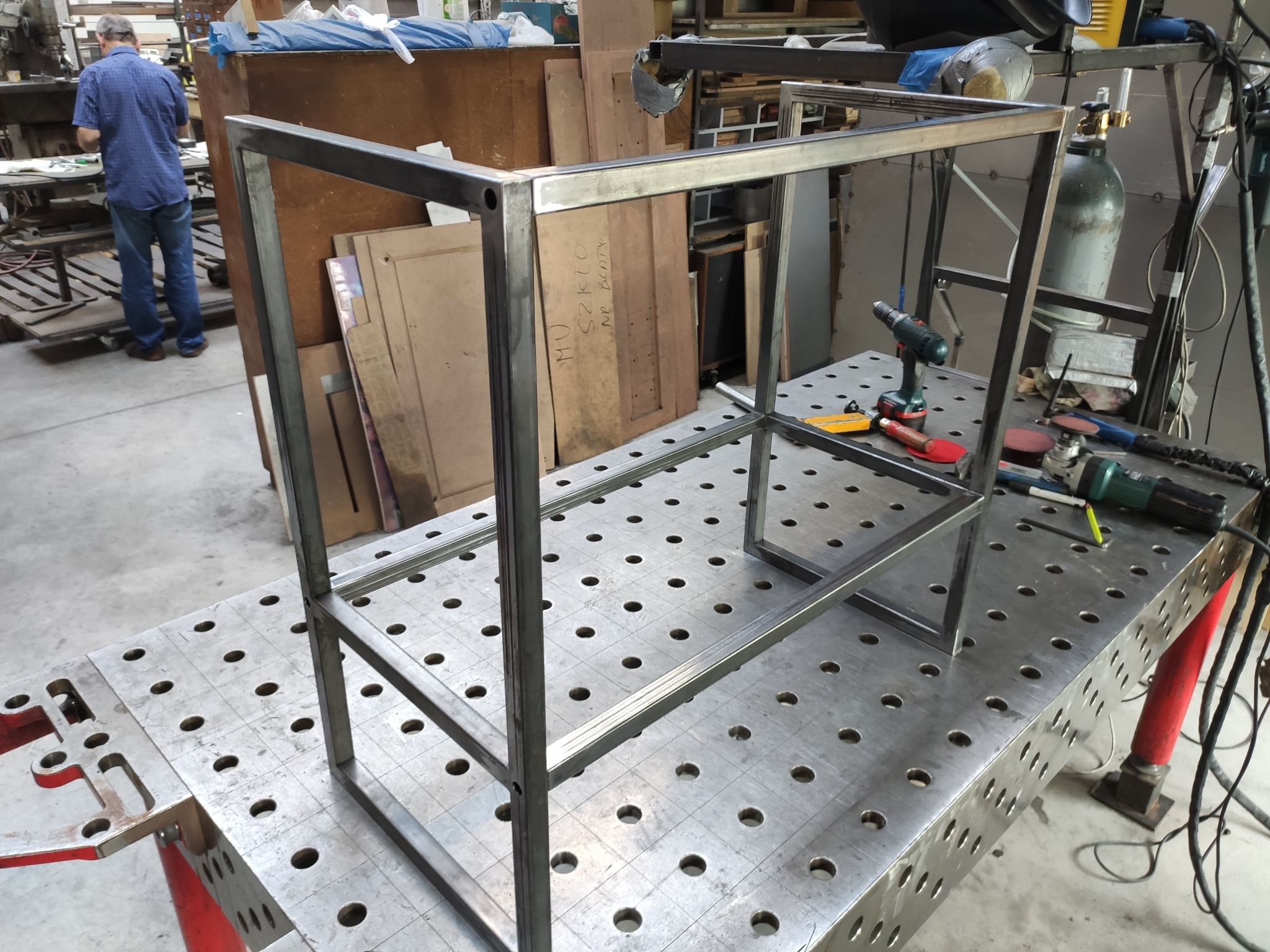 Metalowa konstrukcja spawana na stole spawalniczym z narzędziami, widoczny fragment warsztatu i sylwetka osoby w tle. Konstrukcja w formie prostopadłościanu.