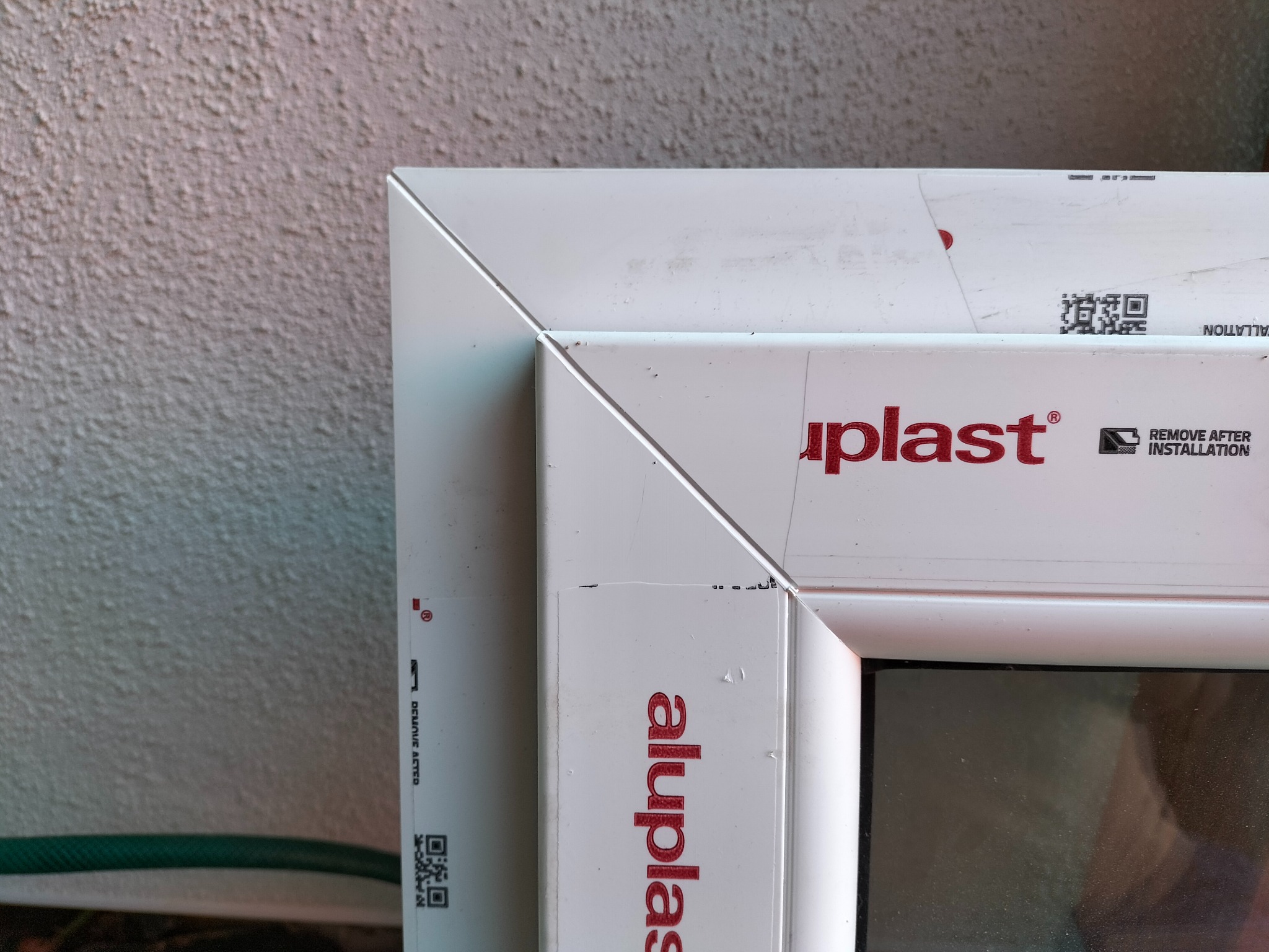 Fragment białej ramy okiennej z tworzywa sztucznego, widoczne logo 'aluplast' i instrukcja usunięcia folii po instalacji, zbliżenie na narożnik i ścianę o chropowatej fakturze w tle.