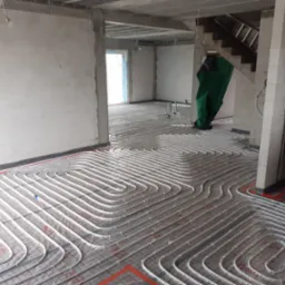 Rozłożona instalacja ogrzewania podłogowego w surowym stanie, białe rurki na czerwonej siatce, widoczne ściany z białego betonu komórkowego, rury PCV w kolorze niebieskim i czerwonym oparte o filar...