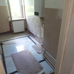 HYDROTECH PW SPÓŁKA Z OGRANICZONĄ ODPOWIEDZIALNOŚCIĄ - Wnętrze w trakcie remontu z zamontowanym piecem gazowym na ścianie, widoczne rury grzewcze, grzejnik pod oknem, fragment podłogi zabezpieczony kartonem, ściany częściowo otynkowane.