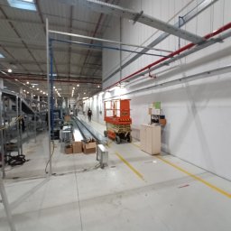 HYDROTECH PW SPÓŁKA Z OGRANICZONĄ ODPOWIEDZIALNOŚCIĄ - Wnętrze hali produkcyjnej z widocznymi instalacjami pod sufitem, pomarańczowym podnośnikiem nożycowym i elementami linii produkcyjnej w tle.