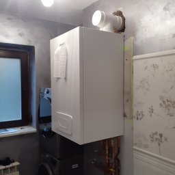 HYDROTECH PW SPÓŁKA Z OGRANICZONĄ ODPOWIEDZIALNOŚCIĄ - Biały piec gazowy zamontowany w łazience, widoczne rury przyłączeniowe, obok pralka, ściana z efektem betonu i fragment tapety w kwiaty.