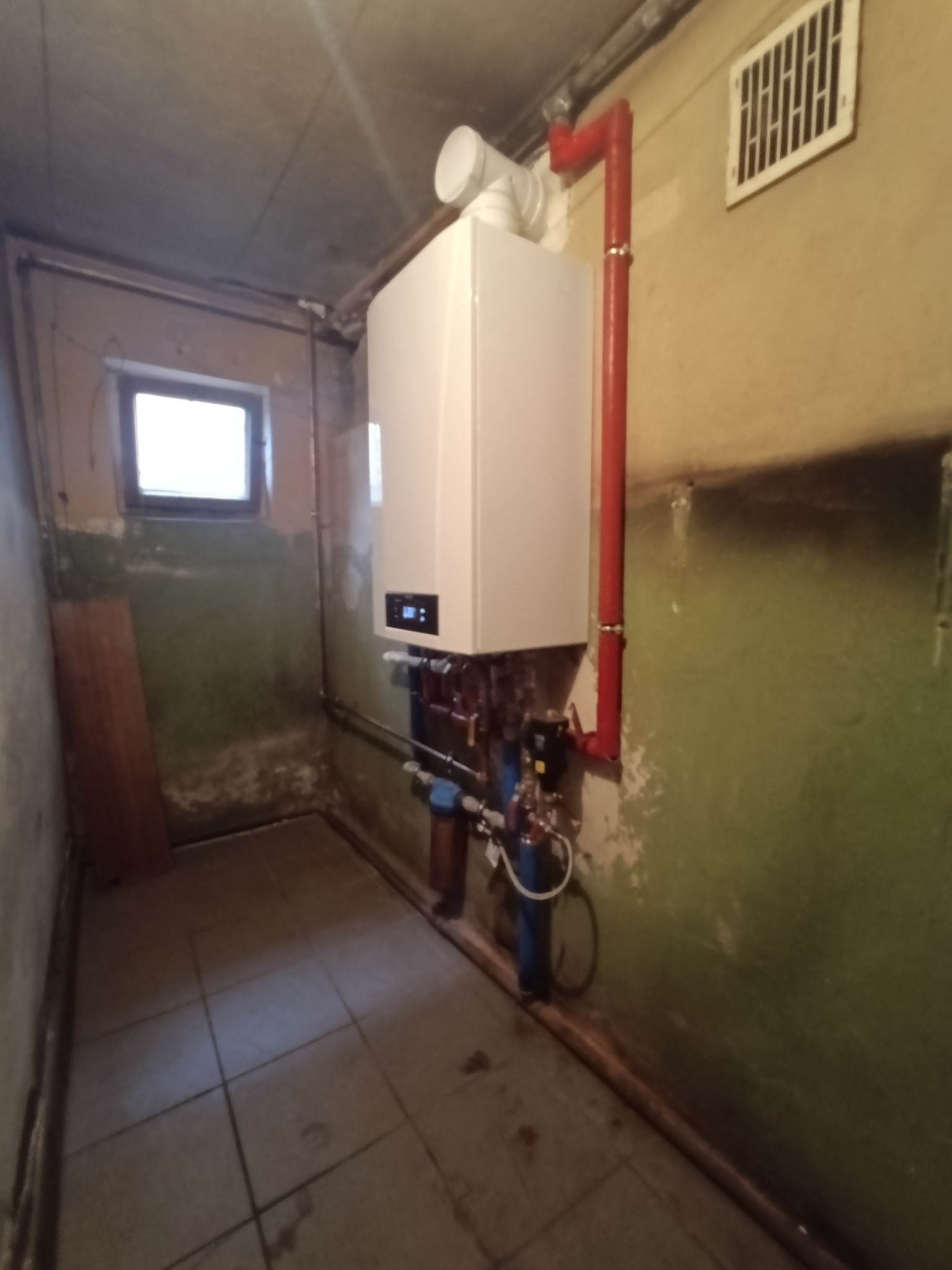 Nowoczesny, biały piec gazowy wiszący na ścianie z widocznymi przyłączami hydraulicznymi i rurą odprowadzającą spaliny, zamontowany w pomieszczeniu z oknem i starą, częściowo pomalowaną ścianą.