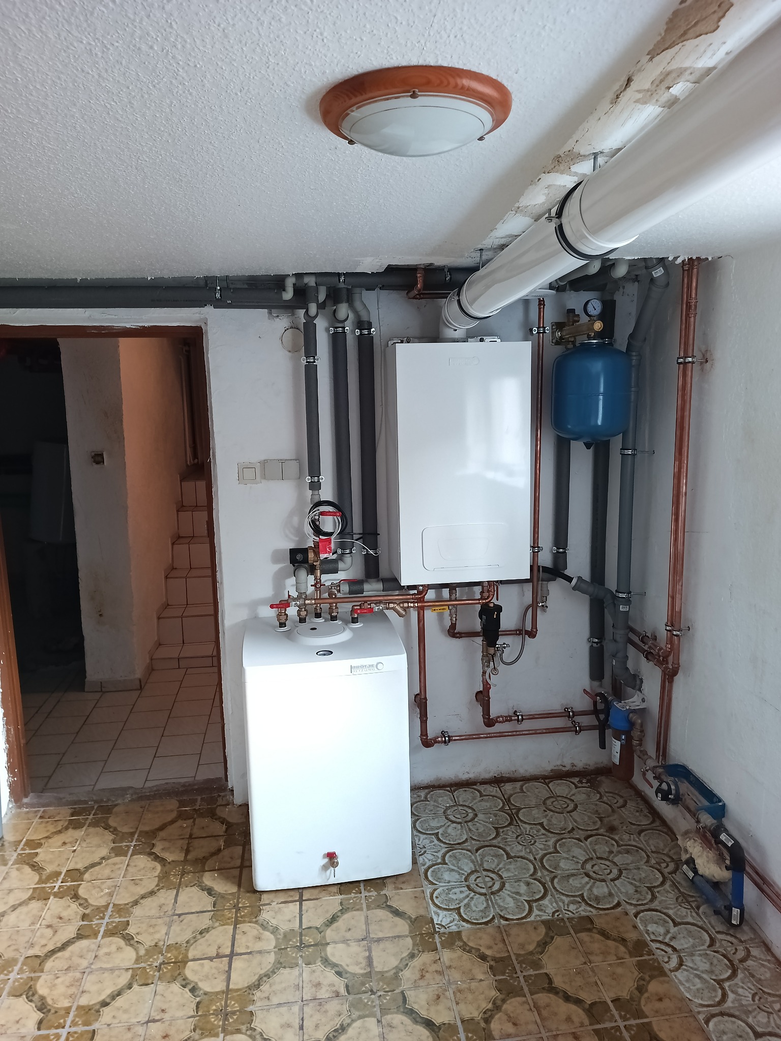 Zestaw dwóch białych pieców gazowych Beretta z niebieskim zbiornikiem wyrównawczym zamontowanych na ścianie, z licznymi rurami miedzianymi i szarymi, w piwnicy z wzorzystą podłogą z płytek.