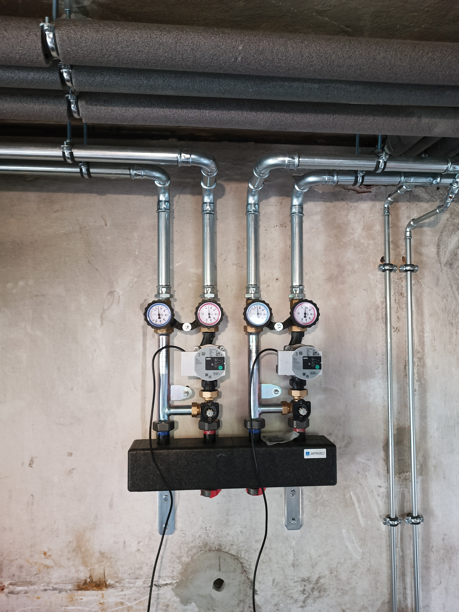 Rozdzielacz instalacji grzewczej z pompami obiegowymi, manometrami i rurami instalacyjnymi zamocowanymi do surowej ściany, widoczne izolowane rury pod stropem.