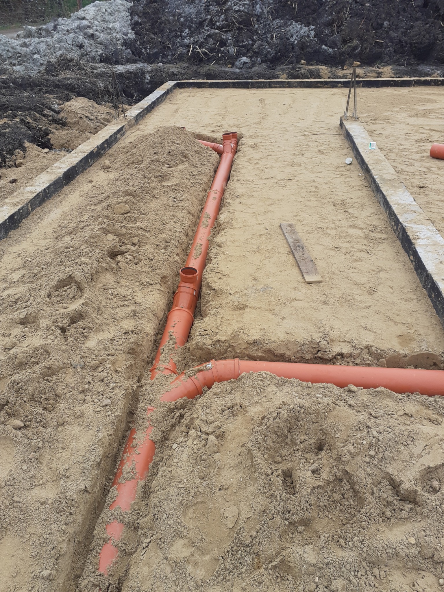 Pomarańczowe rury kanalizacyjne ułożone w wykopie na piaszczystym terenie budowy, widoczne fundamenty i pryzma ziemi w tle.