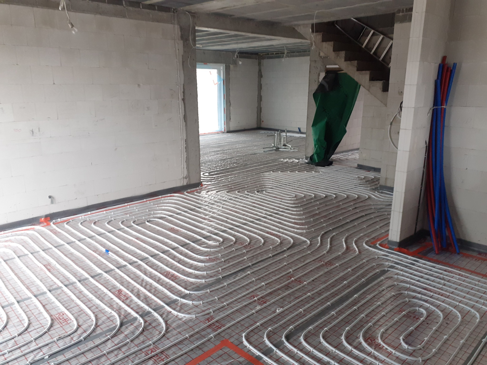 Rozłożona instalacja ogrzewania podłogowego w surowym stanie, białe rurki na czerwonej siatce, widoczne ściany z białego betonu komórkowego, rury PCV w kolorze niebieskim i czerwonym oparte o filar...