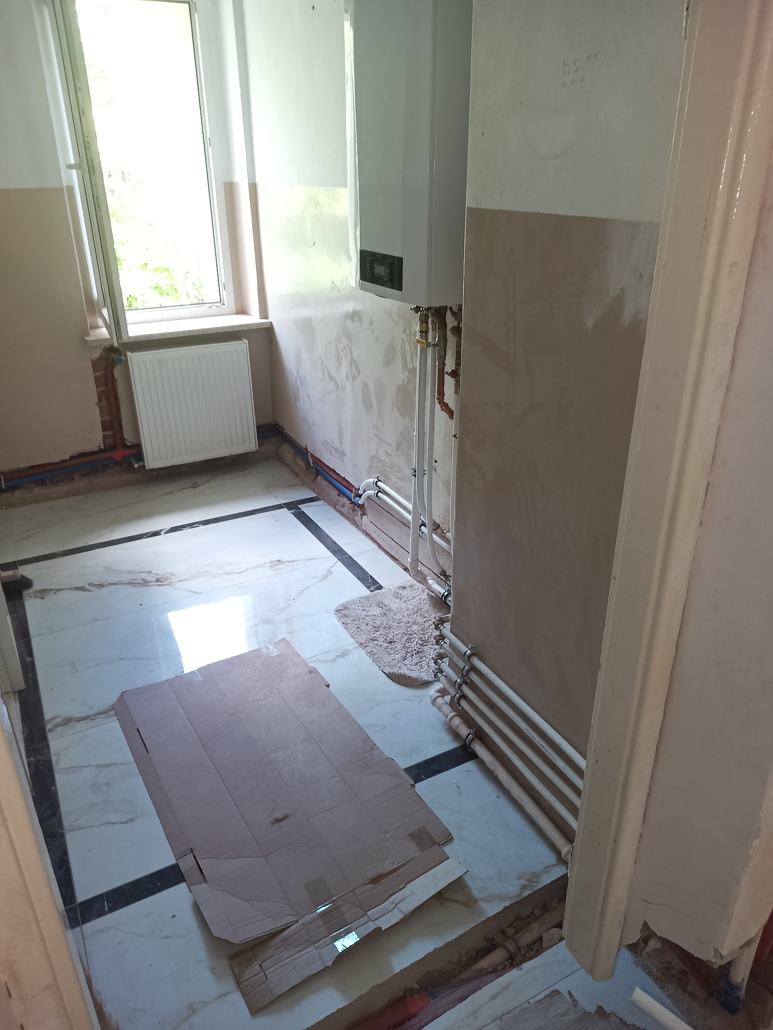 Wnętrze w trakcie remontu z zamontowanym piecem gazowym na ścianie, widoczne rury grzewcze, grzejnik pod oknem, fragment podłogi zabezpieczony kartonem, ściany częściowo otynkowane.