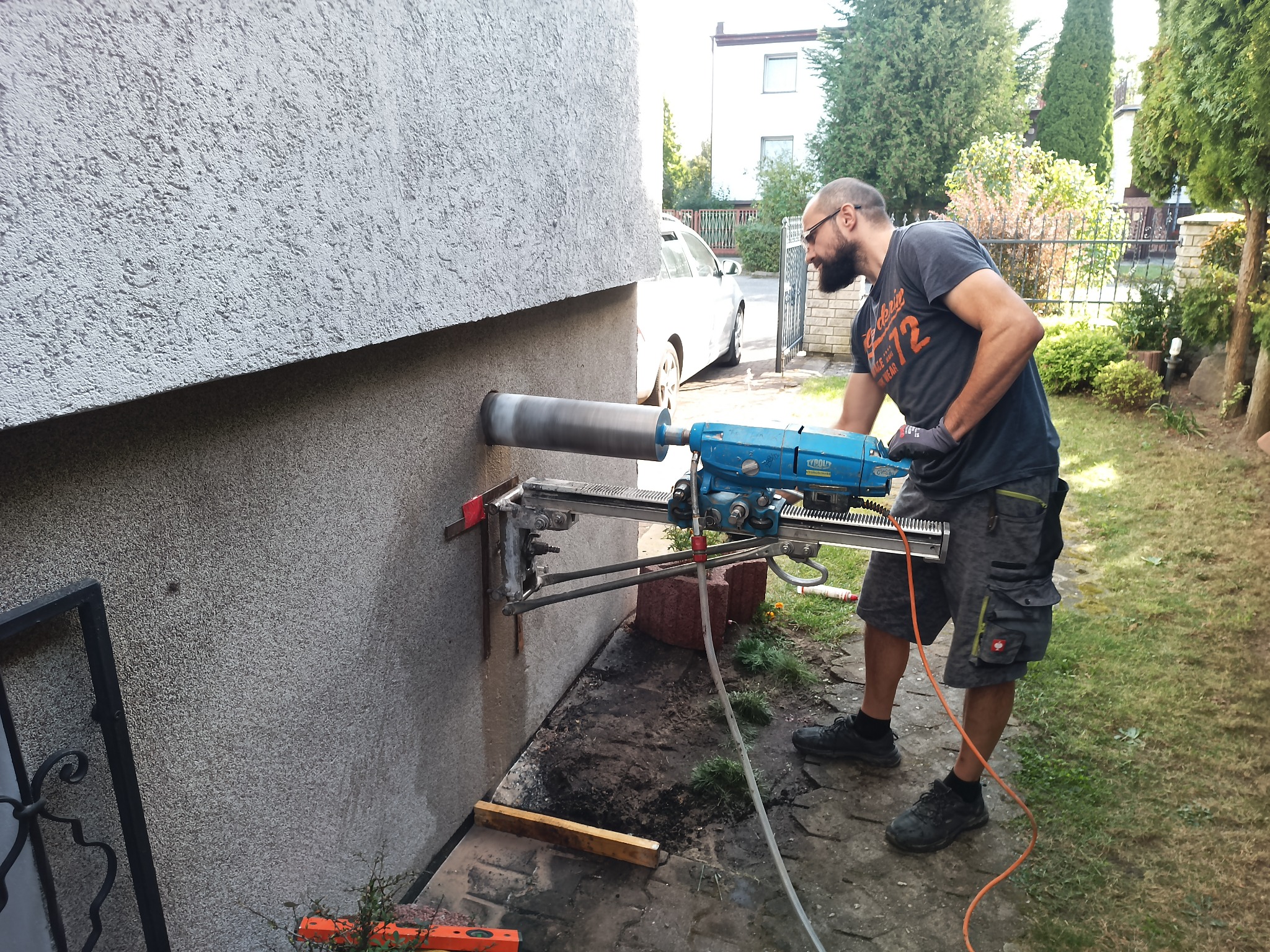 Mężczyzna wierci otwór w ścianie budynku za pomocą wiertnicy diamentowej, widoczny samochód w tle i trawa w otoczeniu.