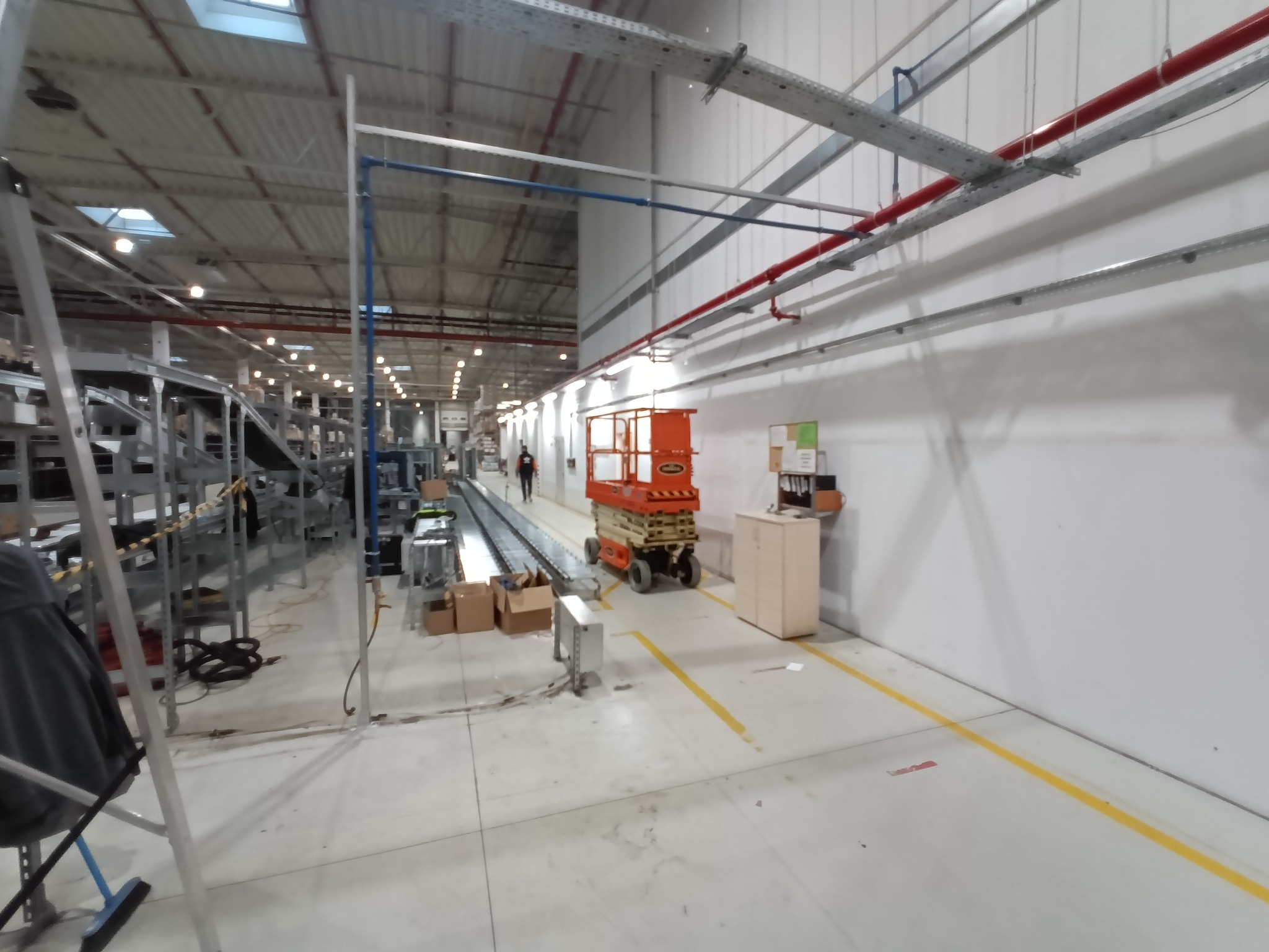 Wnętrze hali produkcyjnej z widocznymi instalacjami pod sufitem, pomarańczowym podnośnikiem nożycowym i elementami linii produkcyjnej w tle.